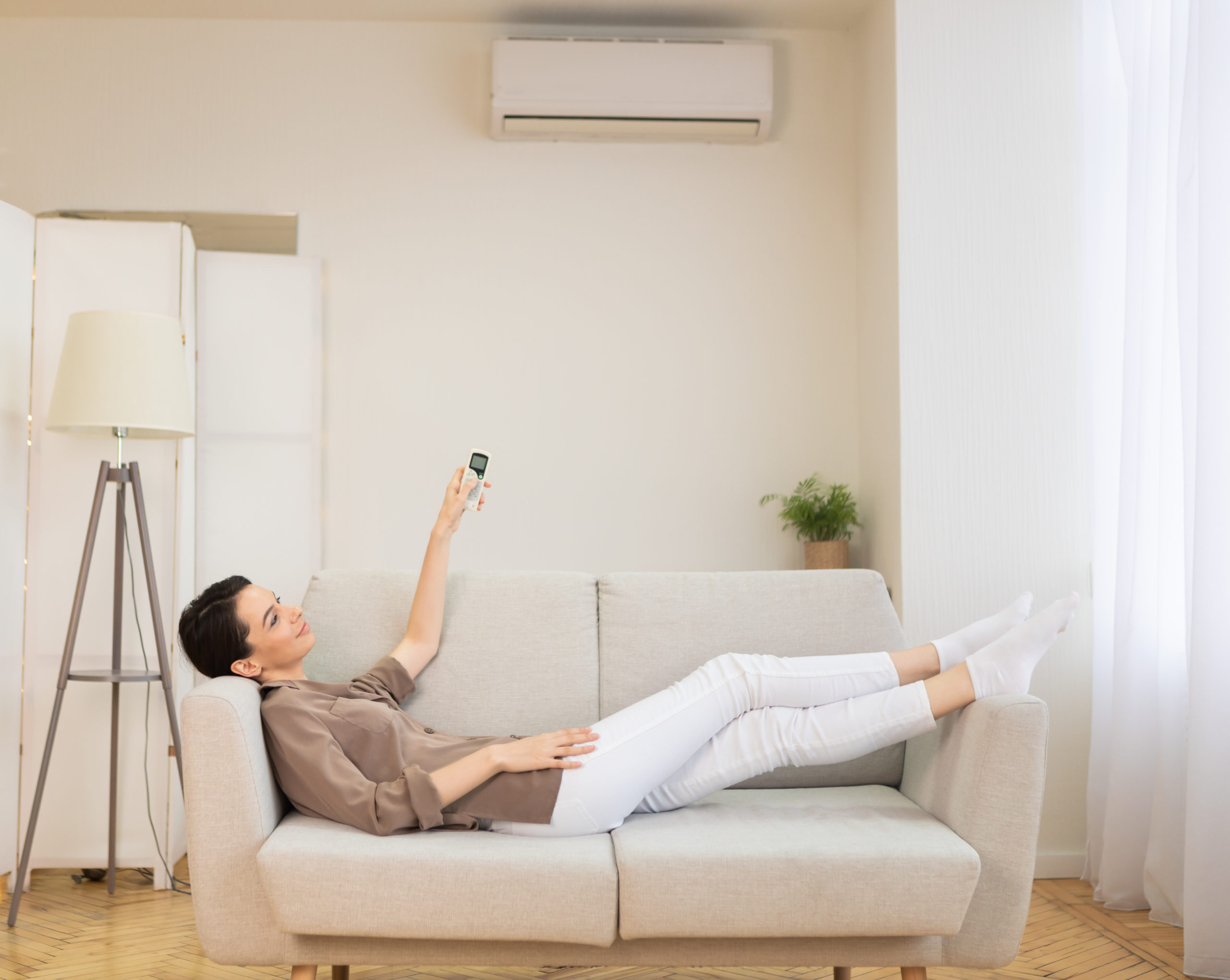 Girl holding remote control relaxing under the air conditioner, lying on couch